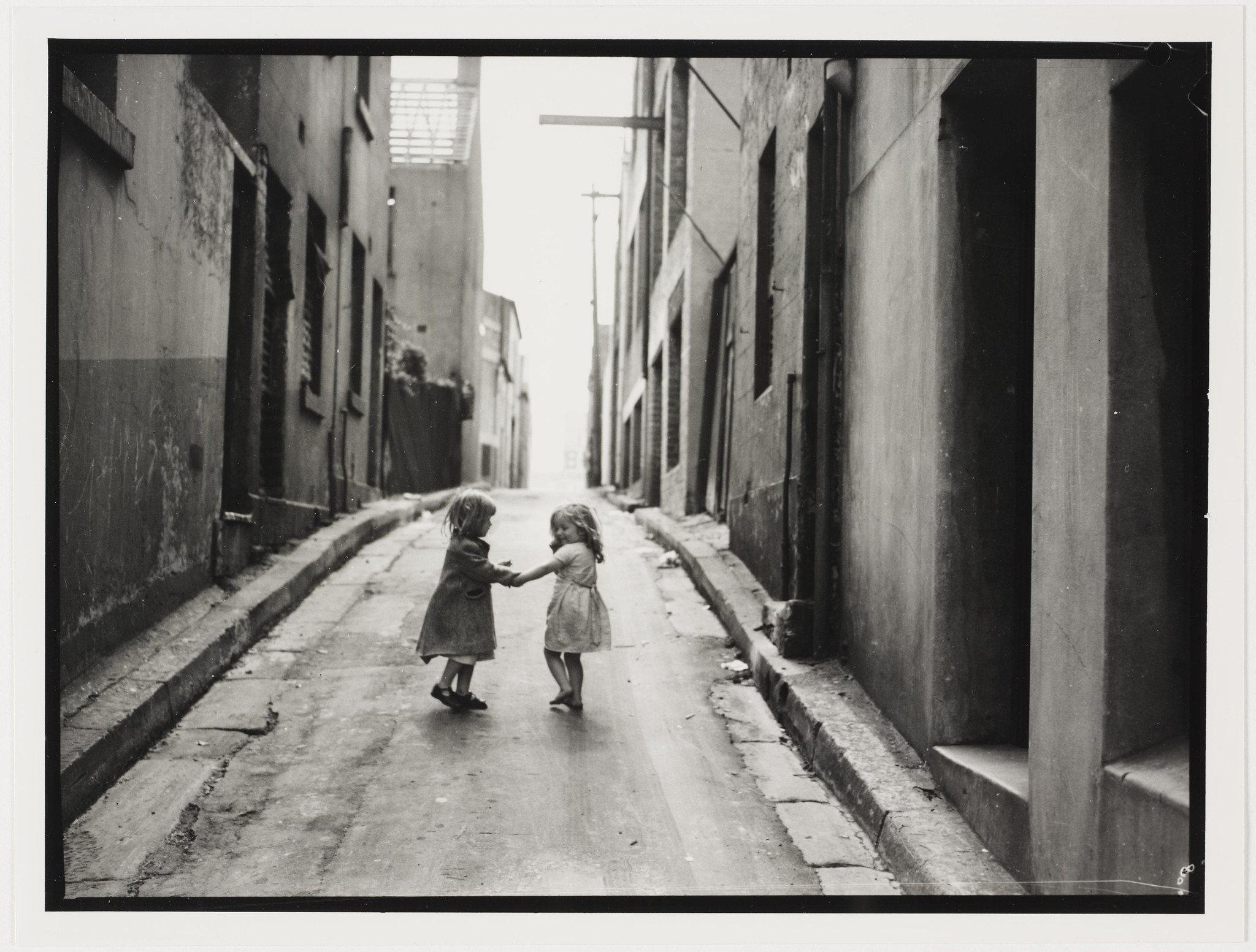 A vintage black and white image of two young girls in dresses holding hands in an alley with talk buildings on either sides. One child is clearly smiling at the other