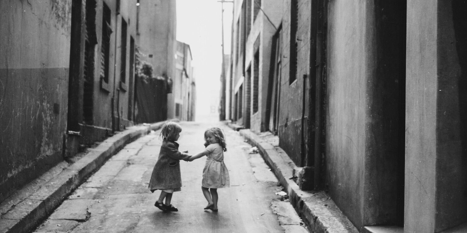 A vintage black and white image of two young girls in dresses holding hands in an alley with talk buildings on either sides. One child is clearly smiling at the other