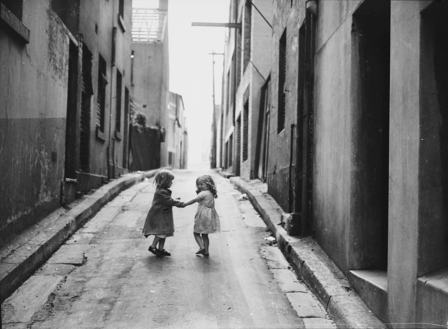 A vintage black and white image of two young girls in dresses holding hands in an alley with talk buildings on either sides. One child is clearly smiling at the other